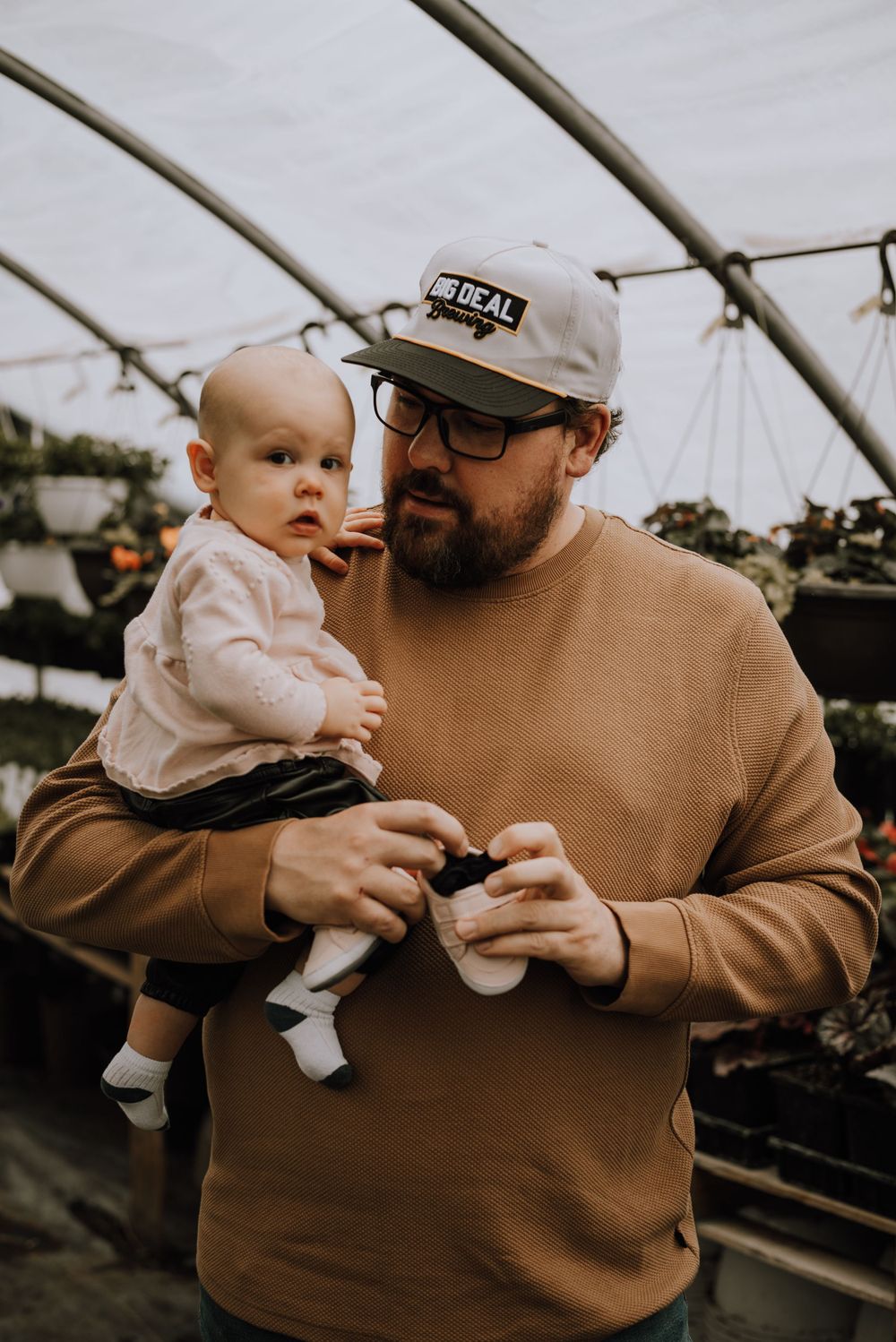 A person in a tan sweatshirt holds a baby in white clothing inside a greenhouse structure.