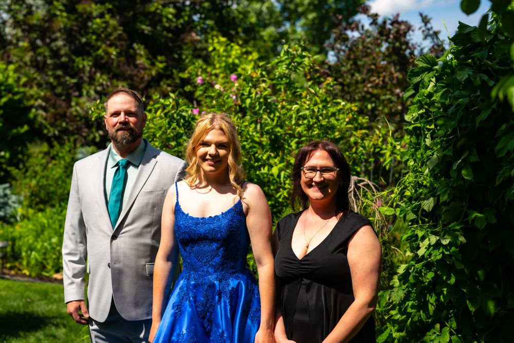 Formal outdoor portrait with three people in elegant attire against green foliage.