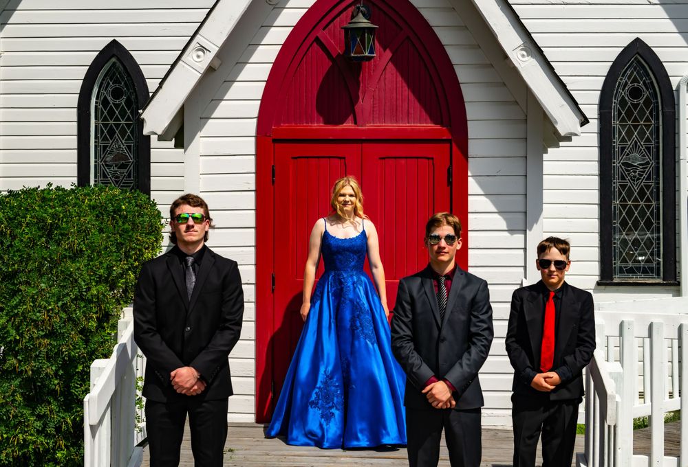Group photo in front of white church with red door, featuring formal attire and blue gown.