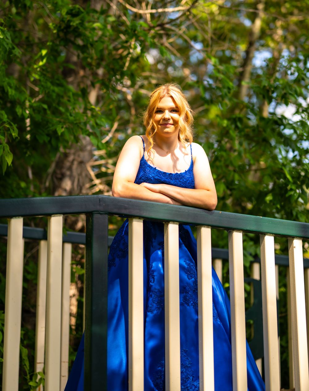 Woman in a royal blue formal dress smiles while standing on a deck railing with lush greenery in the background.