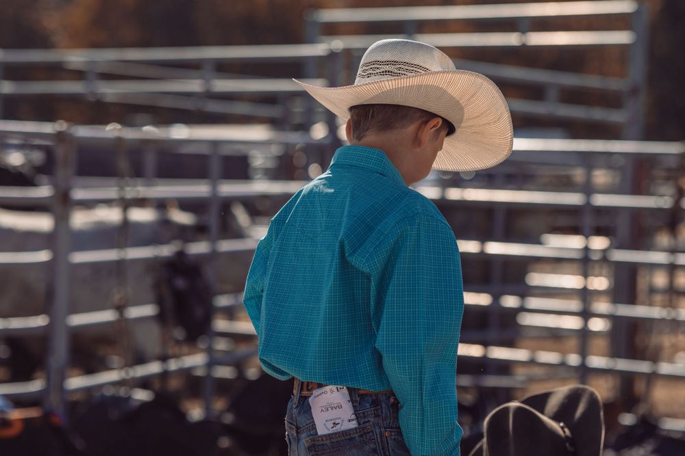 Back view of a young rodeo participant in a turquoise shirt and white cowboy hat near cattle pens.