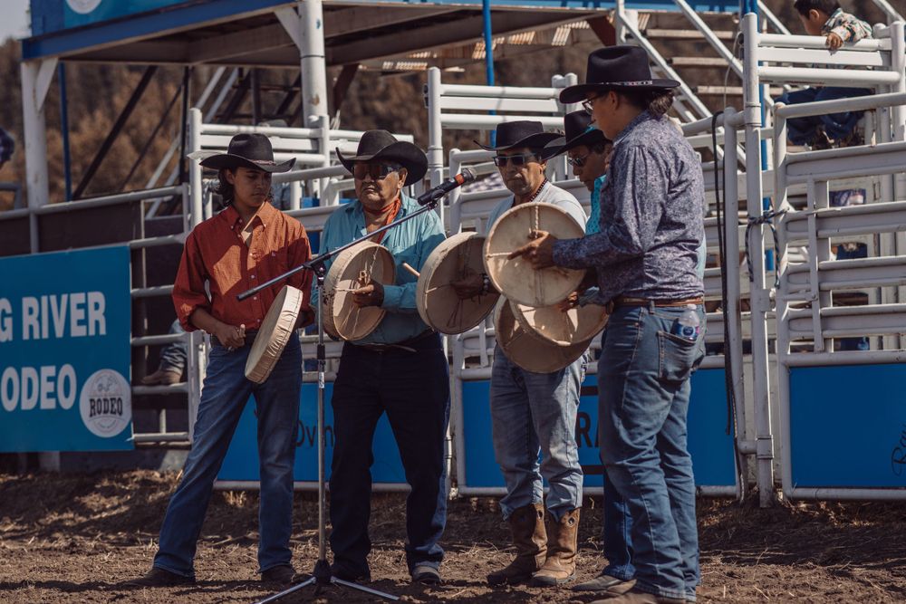 Four cowboys holding large frame drums performing together near rodeo chutes at an outdoor western event.