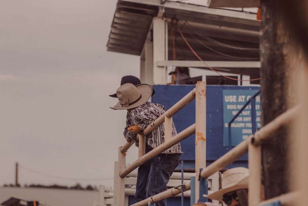 Cowboy in patterned shirt and hat leans over metal fence railing at a rodeo event.