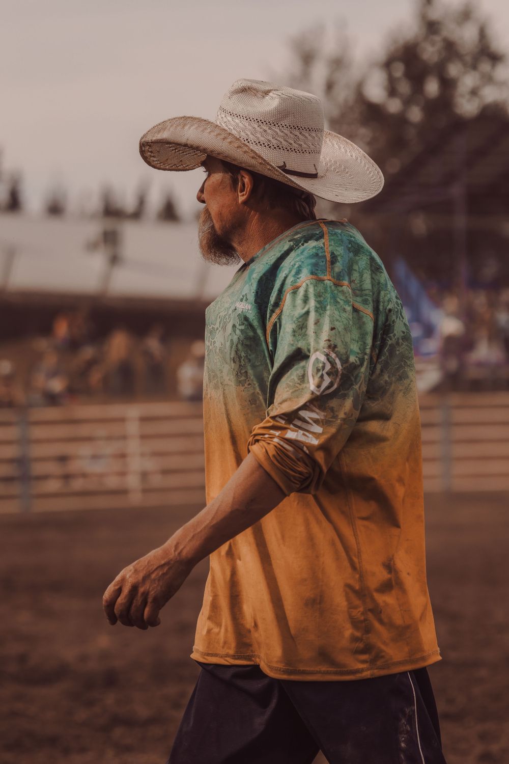 A rodeo clown in a white cowboy hat and colorful tie-dye shirt walks through a dusty arena.