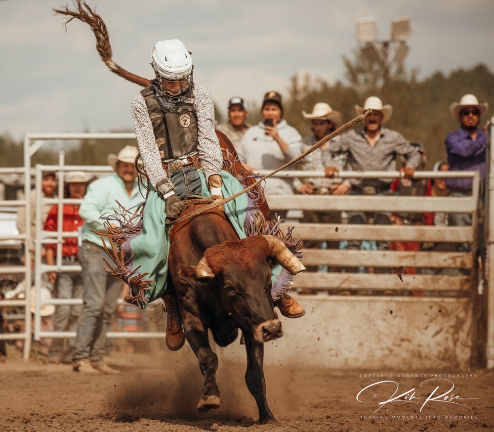 Rodeo competitor in turquoise chaps rides a bucking bull at an outdoor rodeo event with spectators watching.