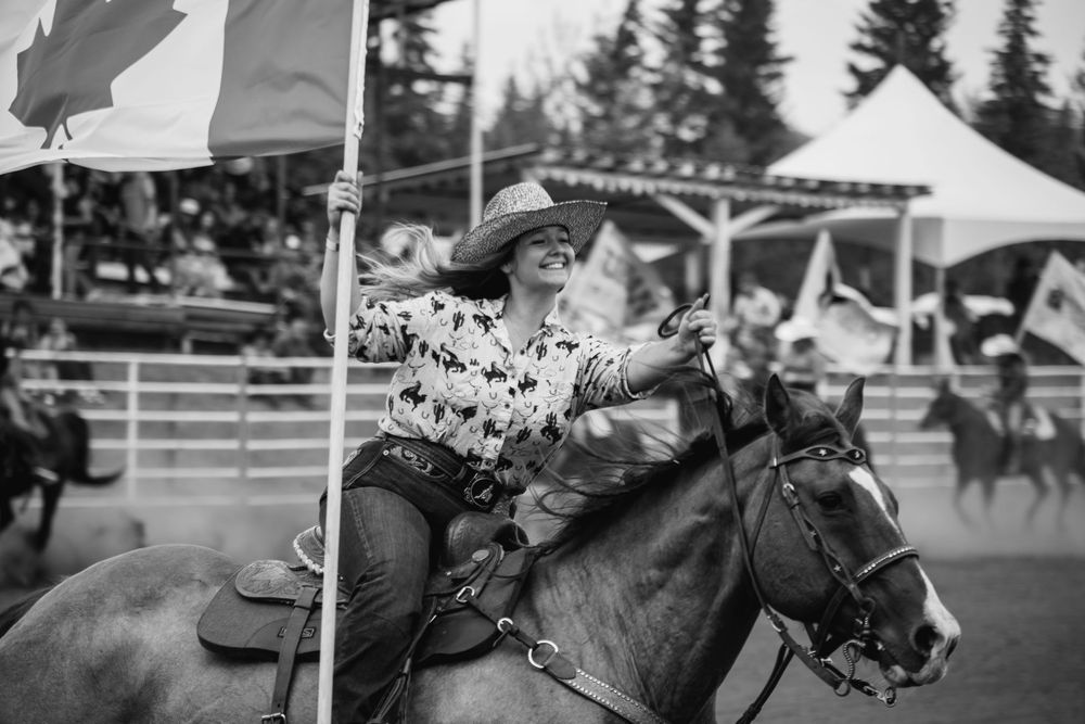 A smiling cowgirl rides a horse while holding a Canadian flag at an outdoor rodeo event in a black and white photo.