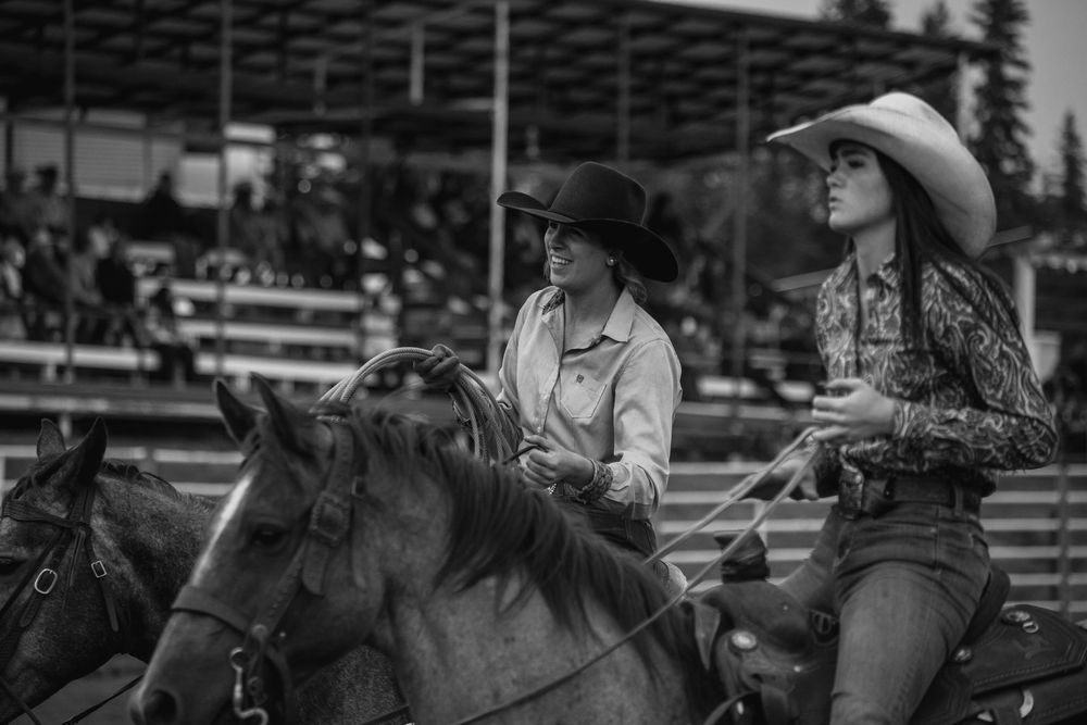 Two women in cowboy hats ride horses at a rodeo arena with spectators in the background.