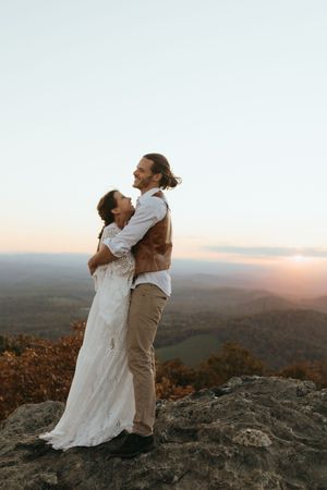A couple shares an intimate moment at sunset on a mountaintop overlook with sweeping valley views in the background.