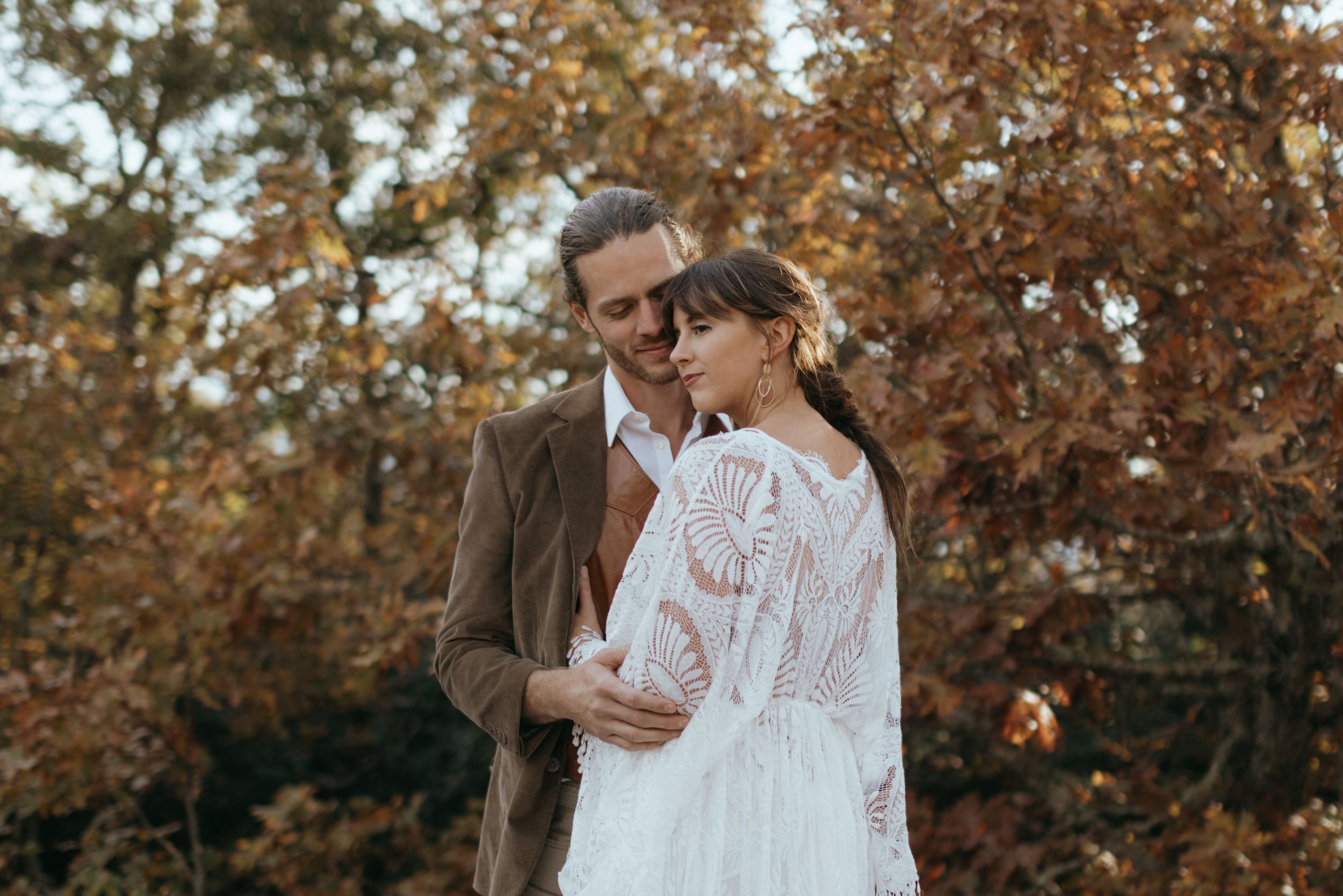 A couple embraces in a romantic autumn setting surrounded by golden-brown leaves and trees.