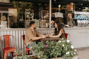 Two people share an intimate conversation at an outdoor cafe with red chairs and decorative plants.