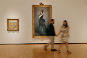 Museum visitors walk past a large framed portrait painting in an art gallery.
