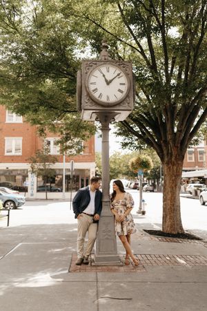 A couple stands beneath a vintage street clock in a quaint downtown setting.