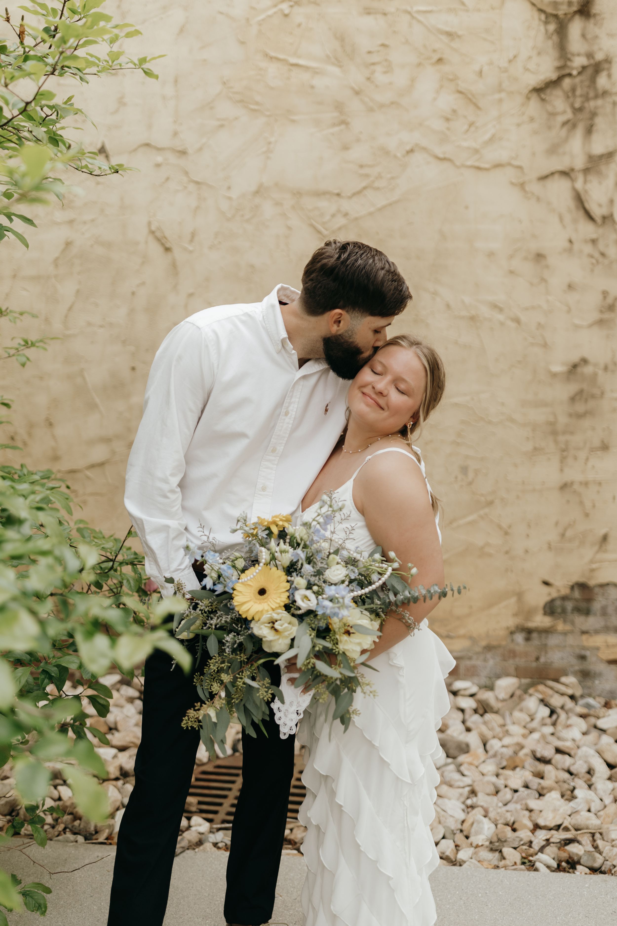 A tender moment between newlyweds with a beautiful wildflower bouquet against a textured wall.