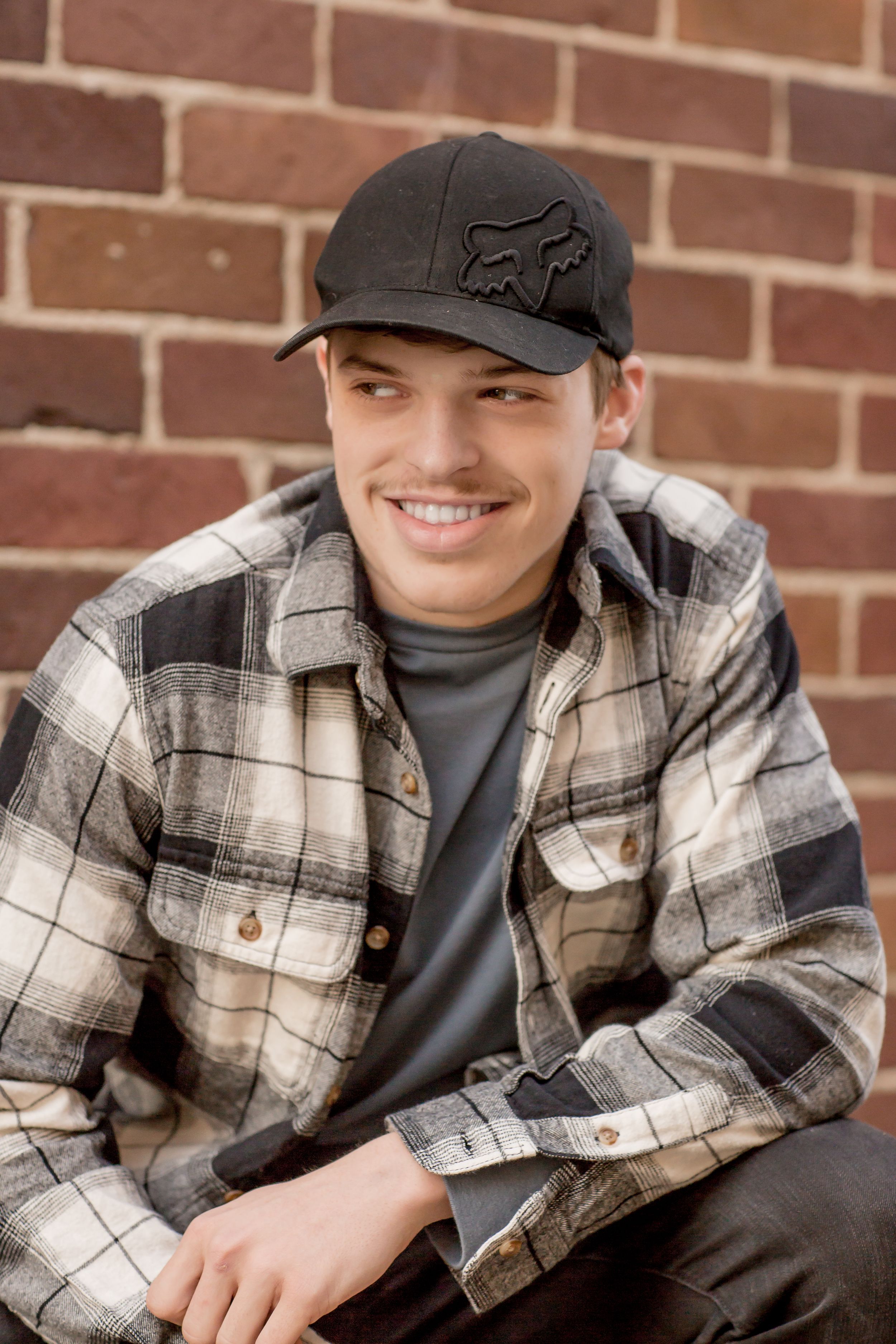 Young man in plaid flannel shirt and black Fox Racing cap smiling against a brick wall.