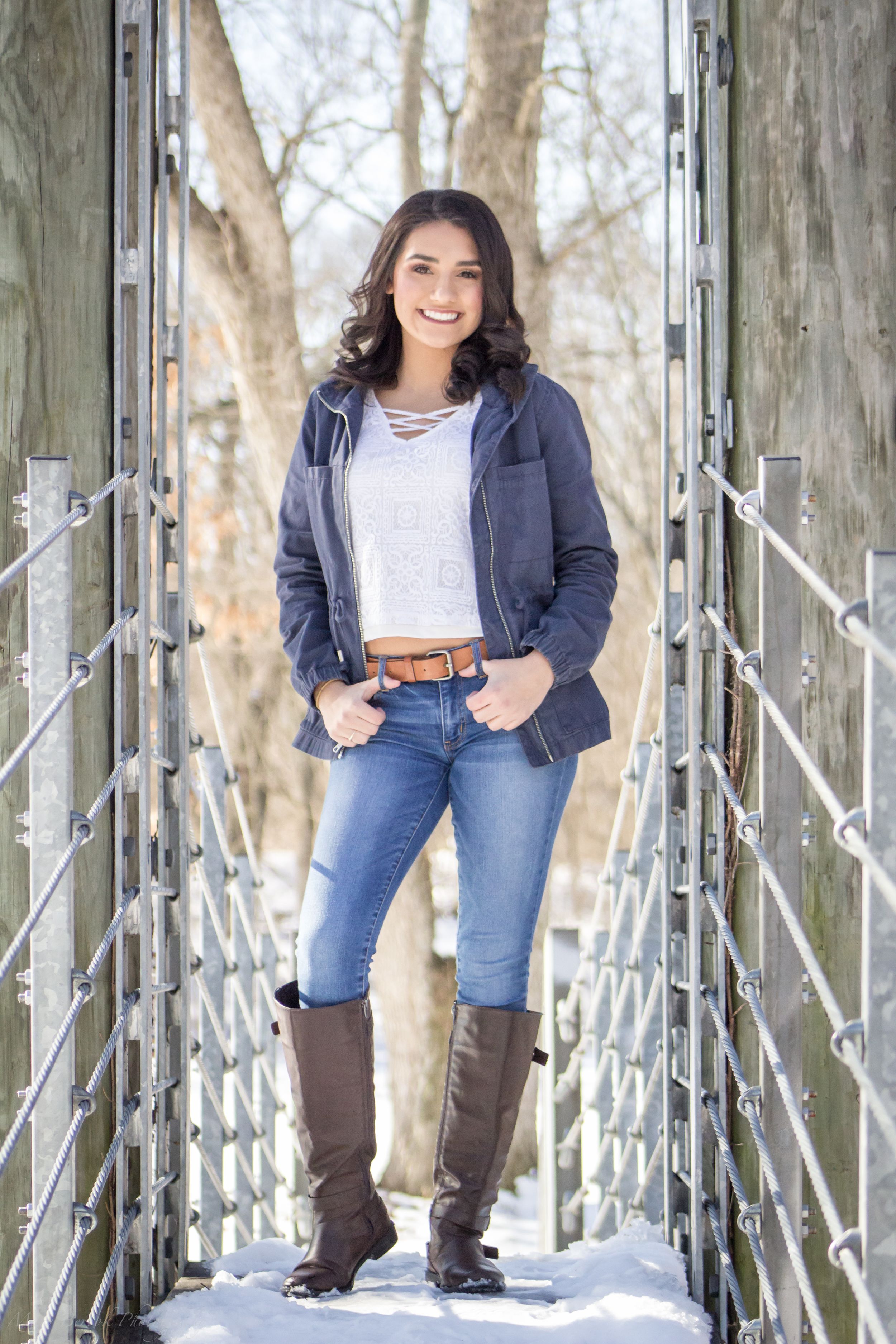 Smiling brunette woman in navy jacket and jeans standing on snowy bridge.