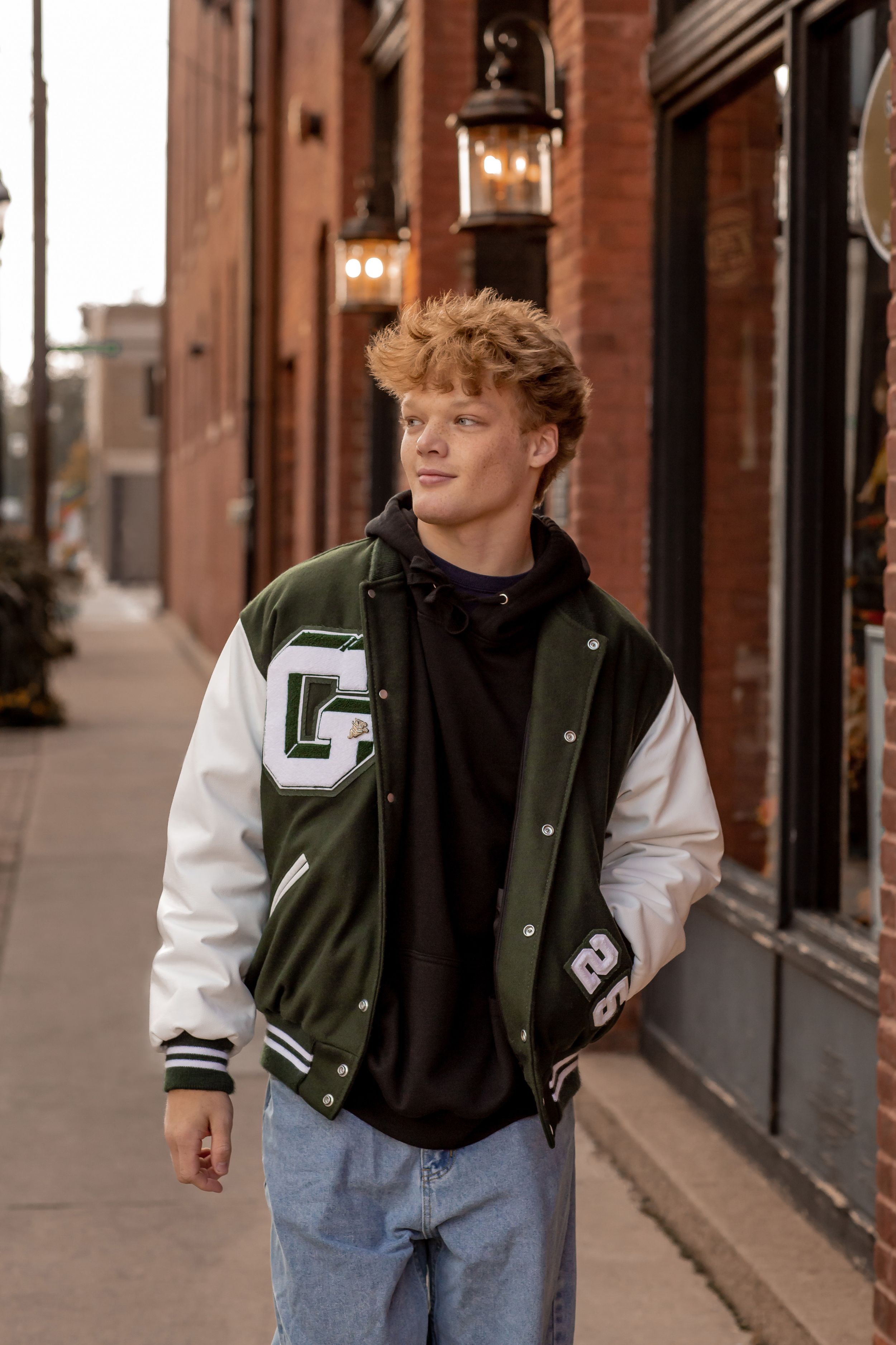 Young man in green varsity jacket walking on urban sidewalk beside brick buildings.