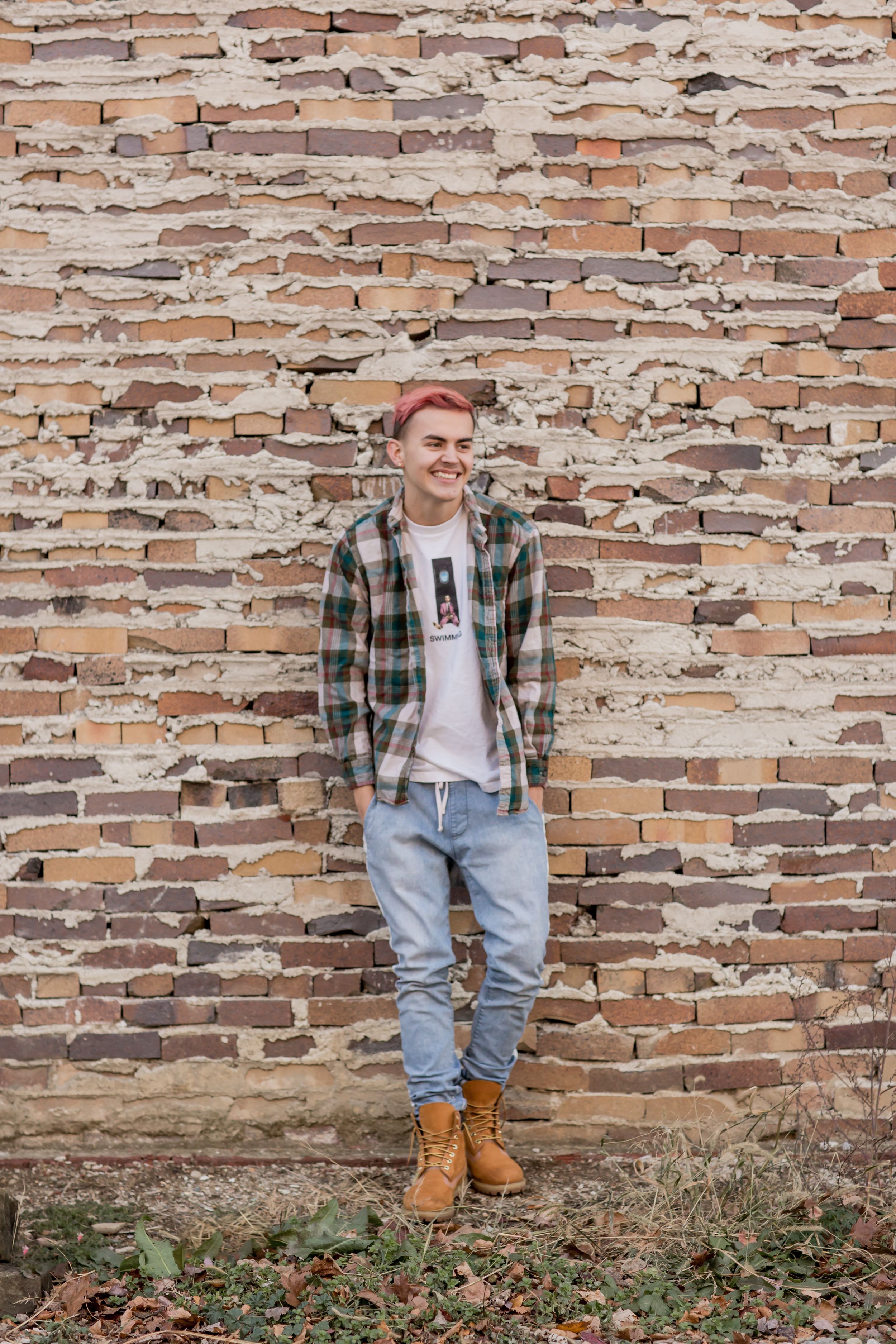 Smiling teen with pink hair in plaid shirt and jeans leaning against rustic brick wall outdoors.