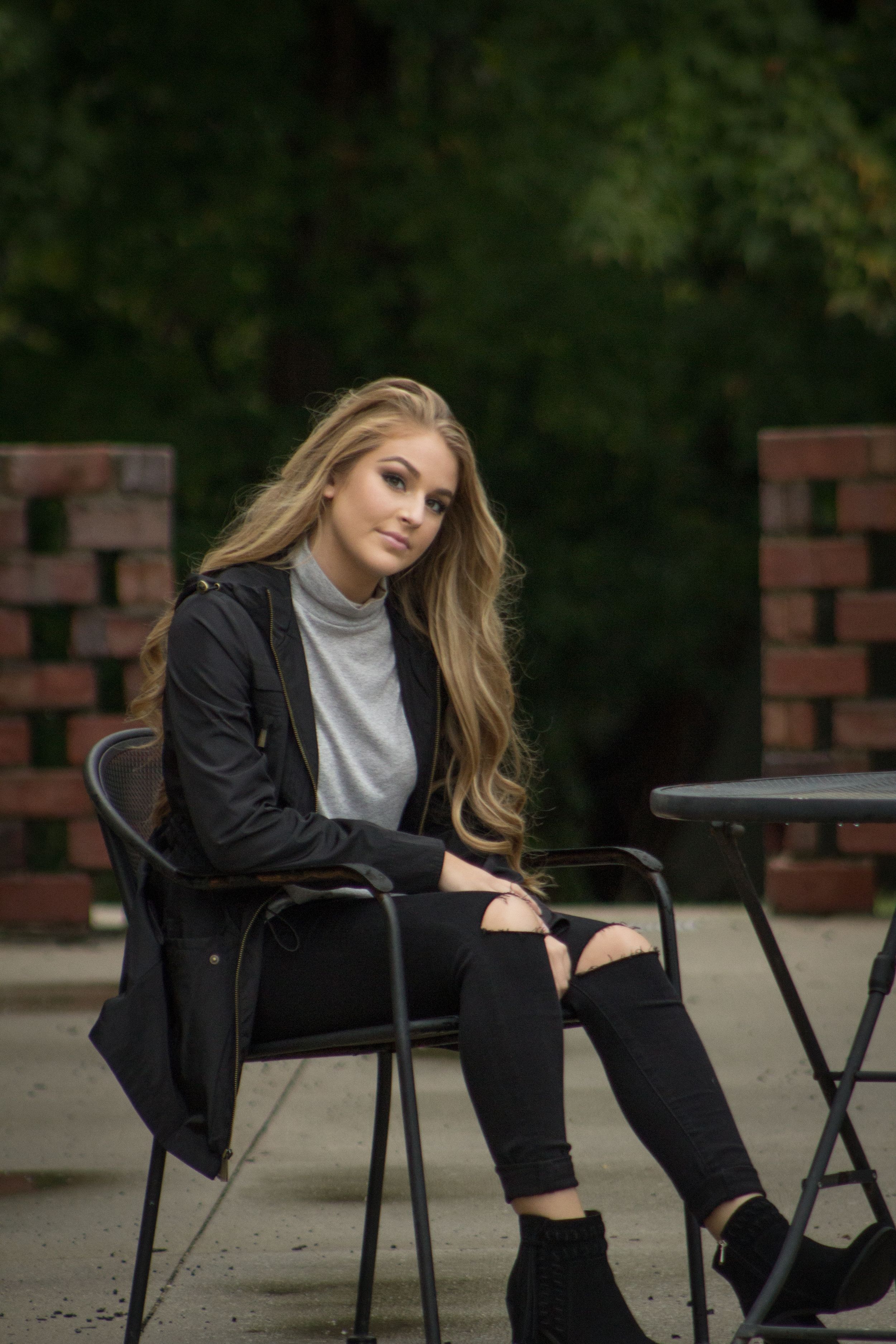 Young woman in black jacket and ripped jeans sitting on chair outdoors near brick wall.