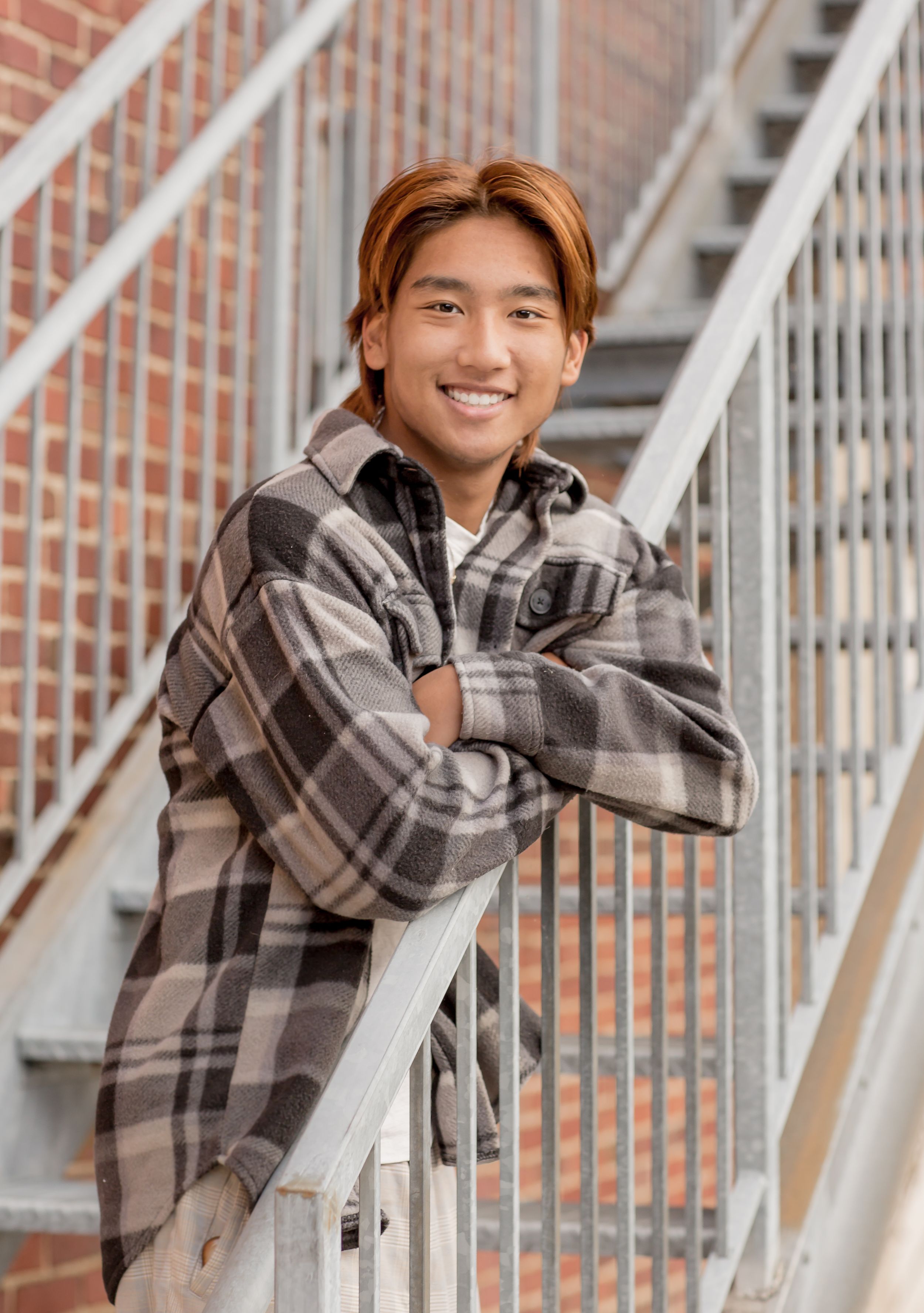 Smiling teen with auburn hair in plaid shirt posing on metal staircase by brick wall.