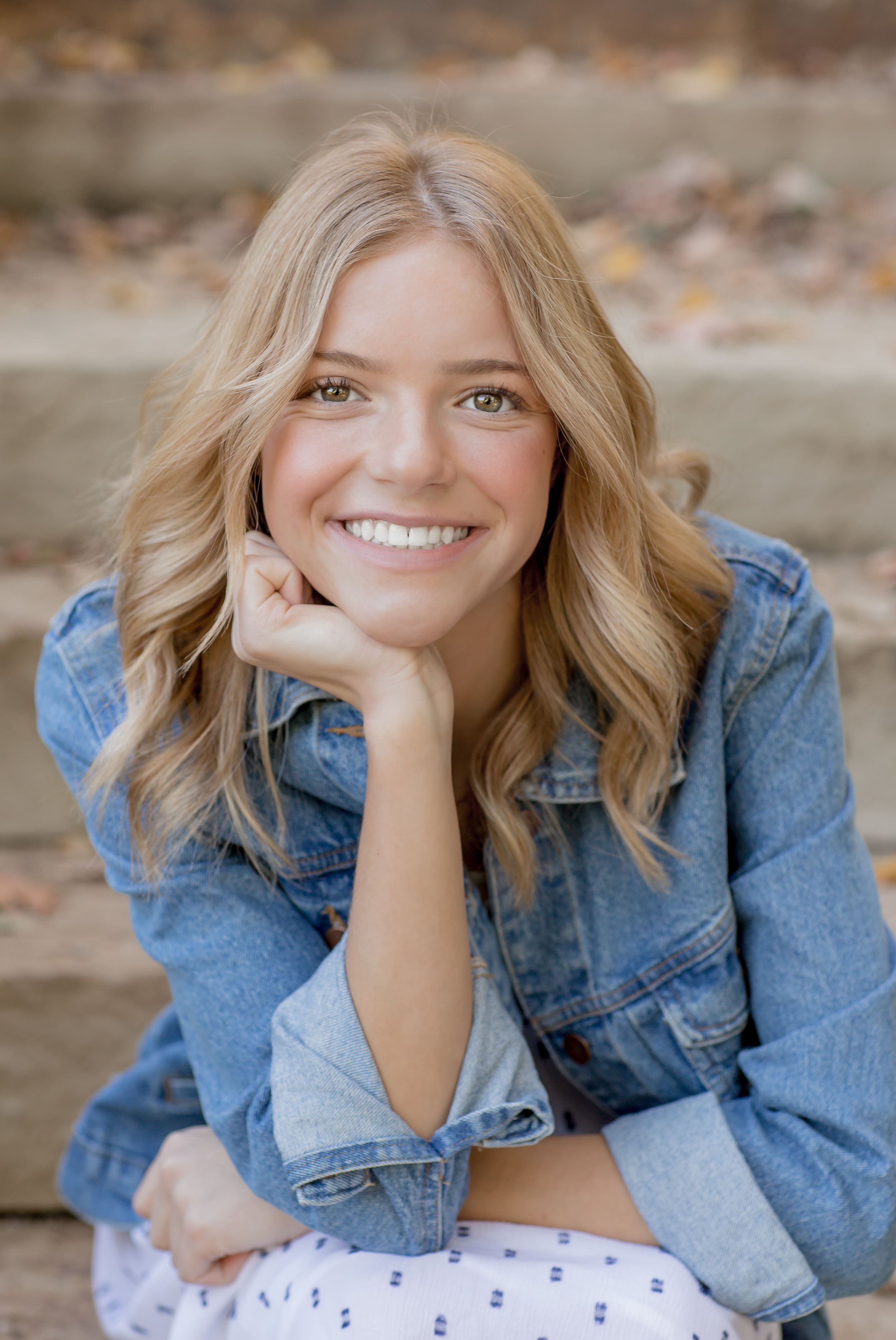 Smiling blonde teenage girl in denim jacket poses on stone steps for fall senior portrait.