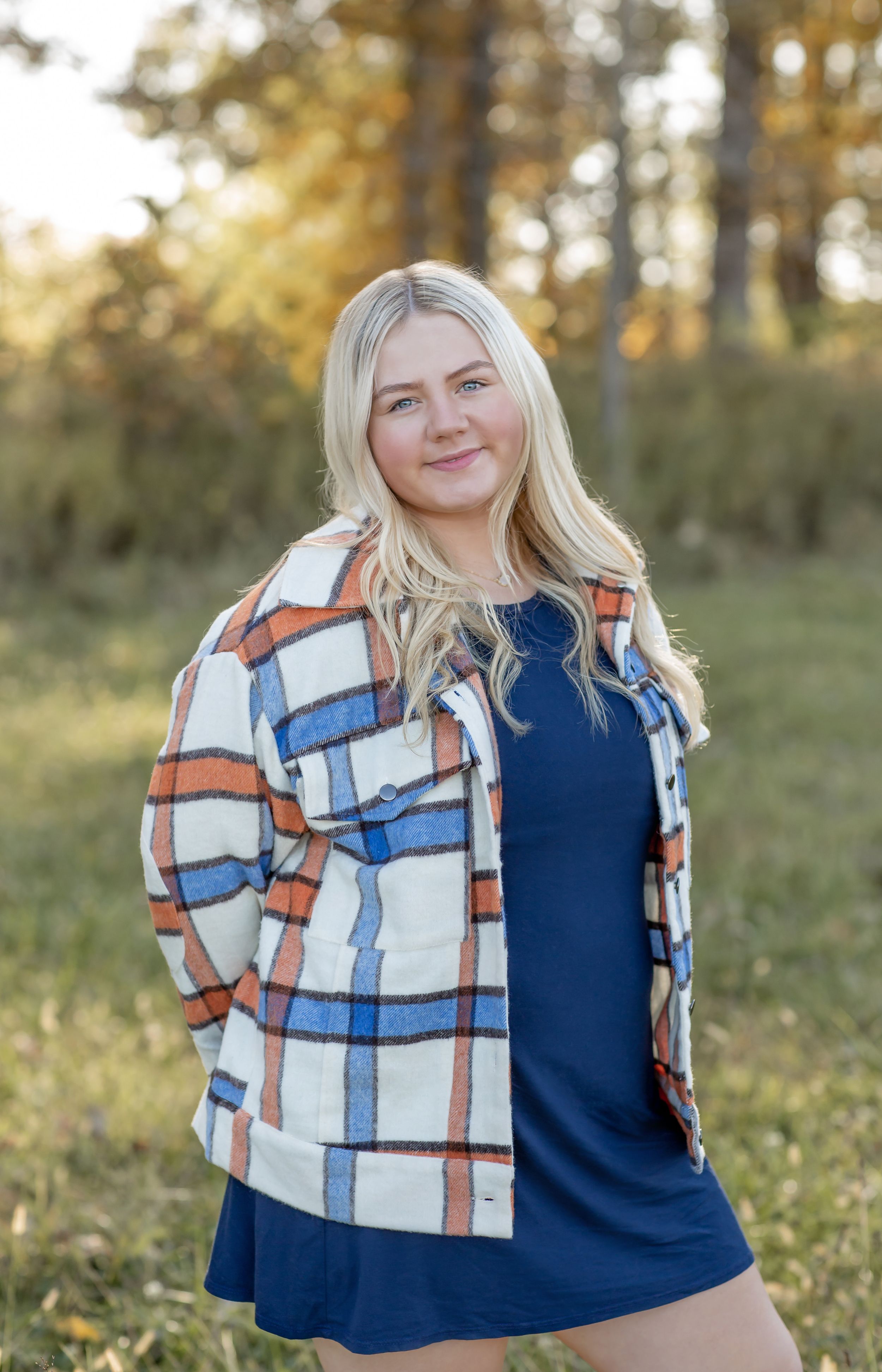 Blonde woman in plaid jacket and navy dress posing outdoors in an autumn forest setting.