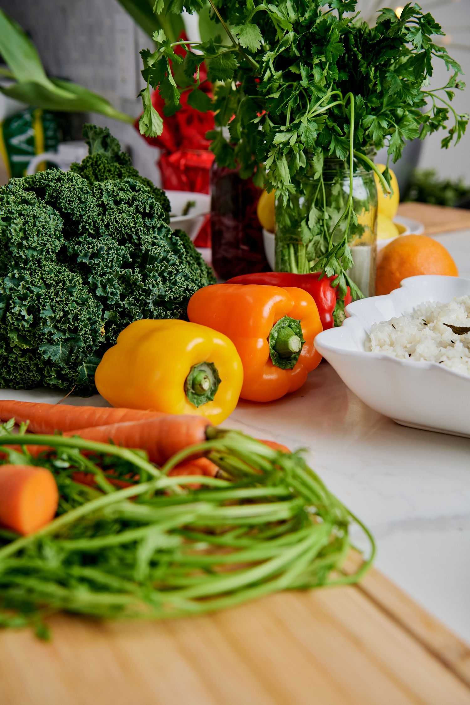 Fresh vegetables including kale, orange bell peppers, carrots and herbs arranged on a wooden cutting board with white bowl.