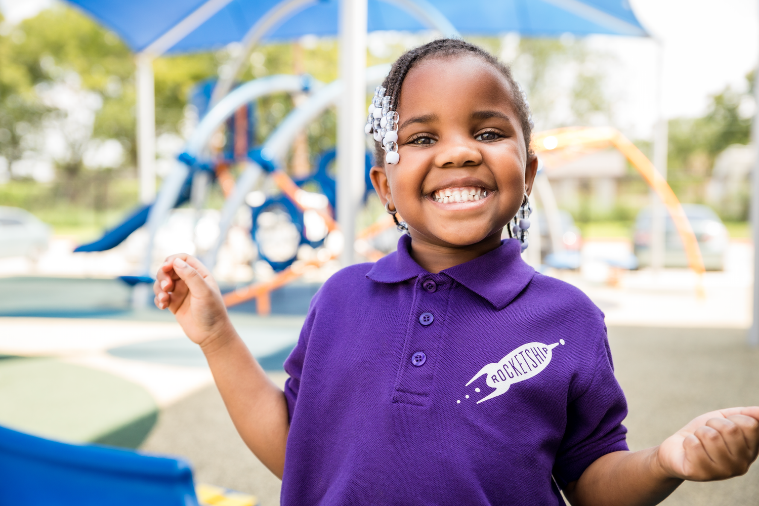 Children in purple polo shirts play and smile at an outdoor playground with blue equipment.