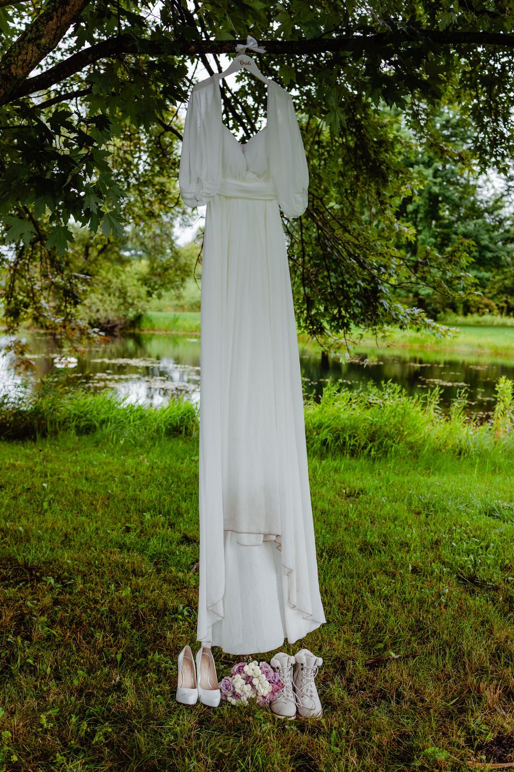 White wedding dress hanging from tree branch over pond with shoes beneath.