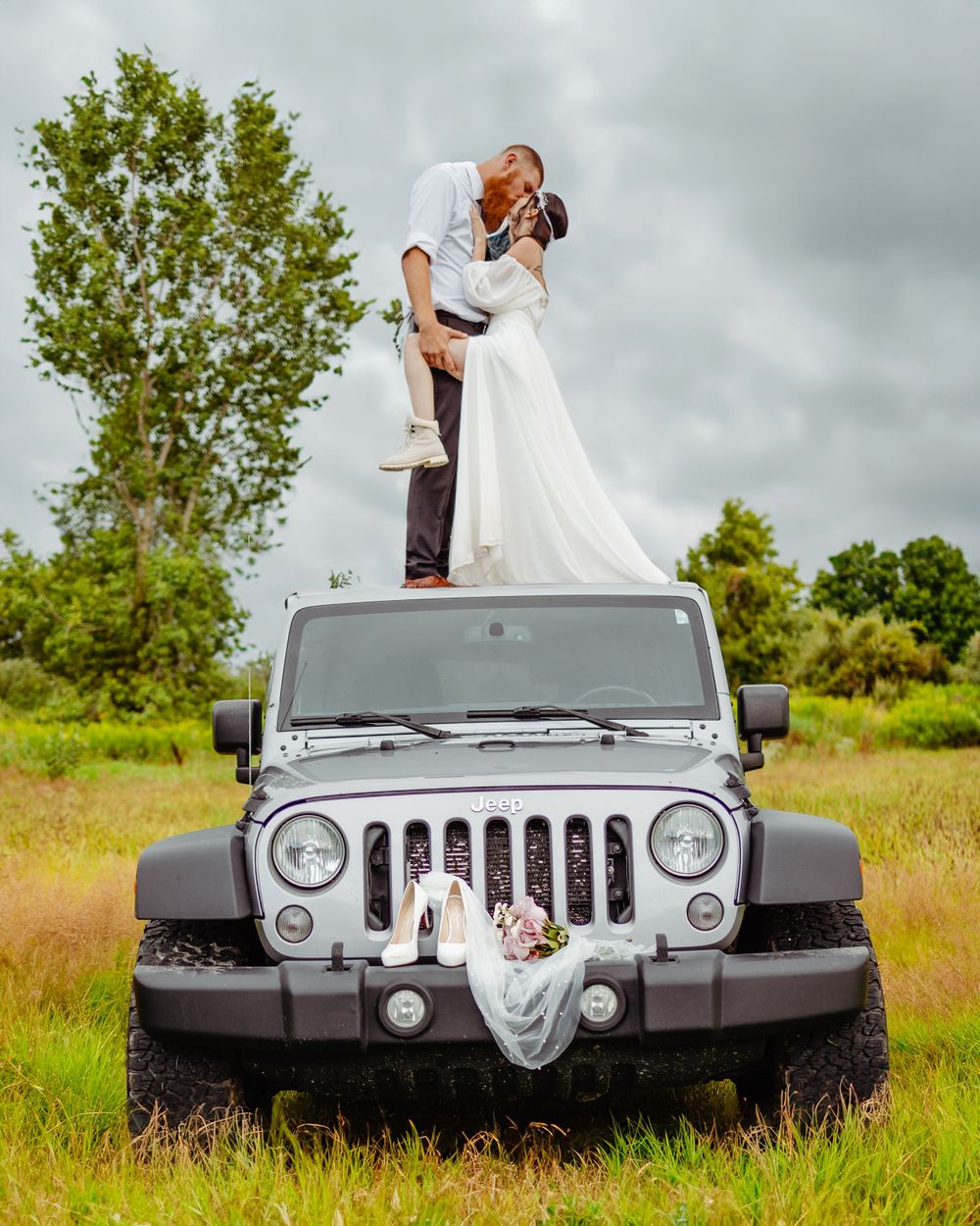 Wedding couple posing dramatically on silver Jeep Wrangler in rural setting.