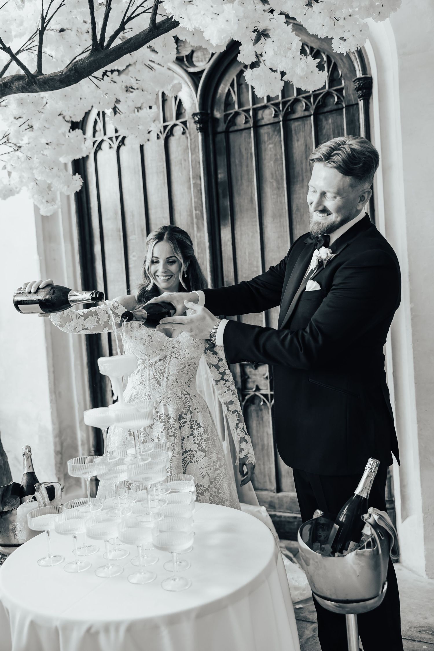Black and white photo of a groom pouring champagne tower at wedding reception at Ashridge house while bride smiles in lace gown.