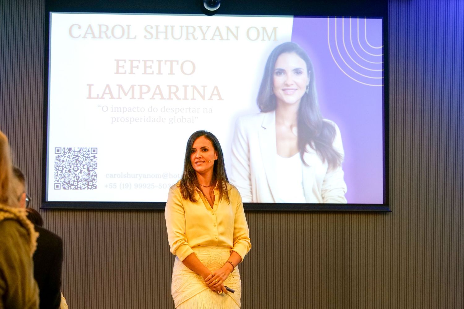 Carol Shuryan OM standing before Efeito Lamparina presentation screen at business conference.