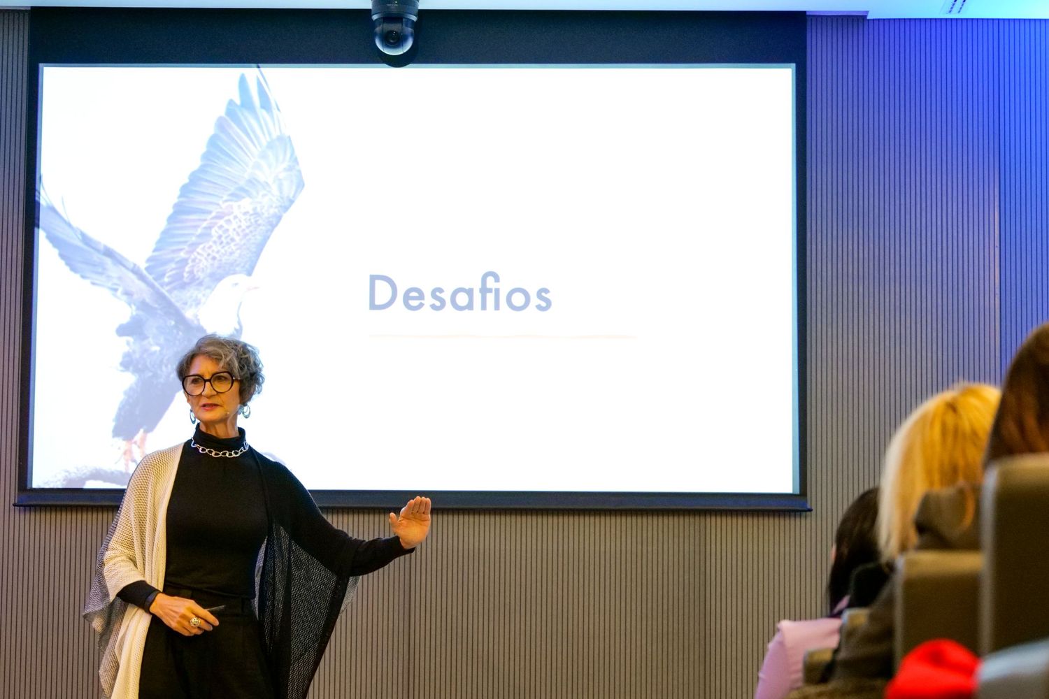 Woman presenter speaks on stage before a screen displaying Desafios during a professional conference.