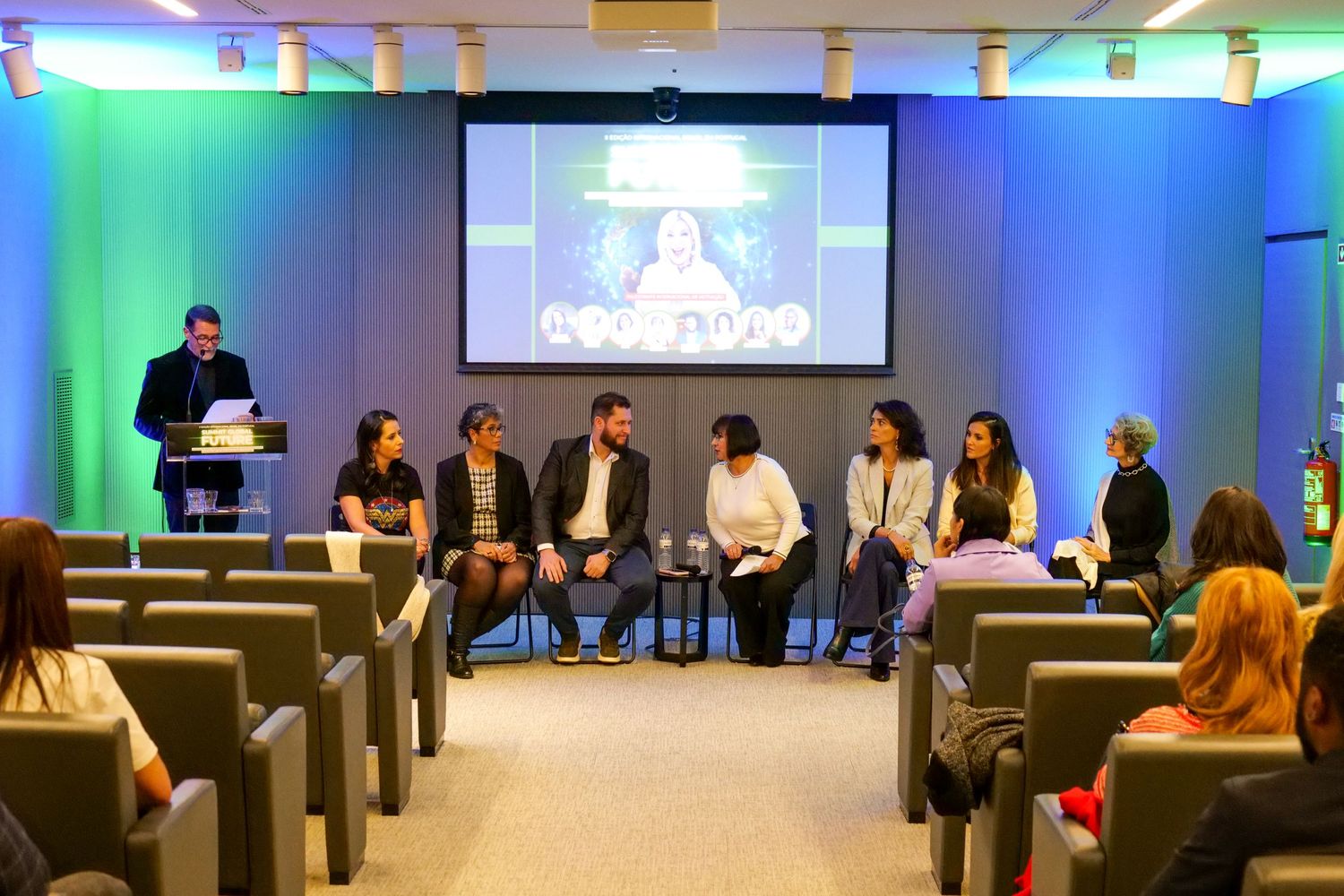 Panel of seven speakers seated on stage during a professional conference in a modern auditorium.