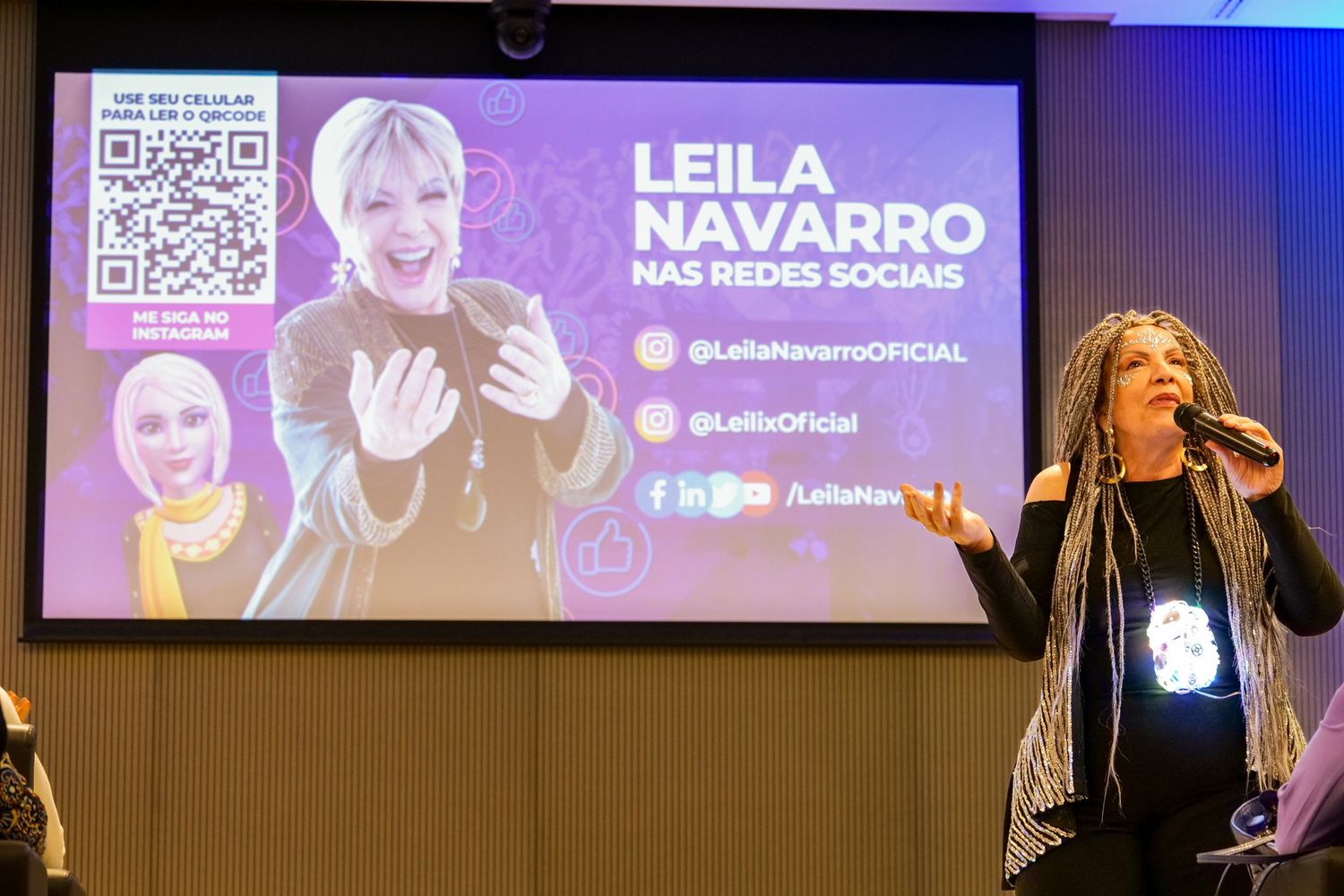 Leila Navarro speaks on stage before a promotional social media screen at a live event.