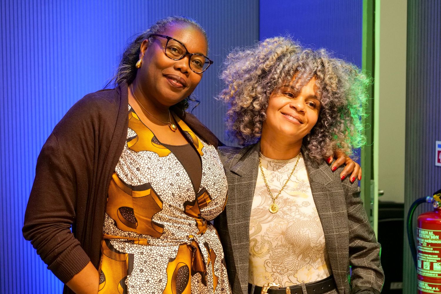 Two women smile warmly together at an indoor professional event.