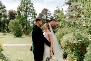 A couple in formal wedding attire embrace while standing in a lush garden setting.