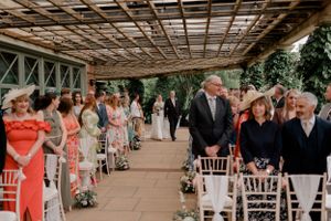 Wedding ceremony guests seated under a latticed pergola decorated with floral arrangements.