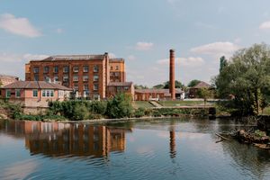 Brick industrial building with tall chimney reflects in calm river water on sunny day with blue sky and trees.