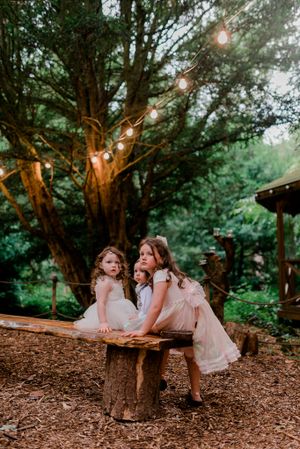 Children in pink dresses sitting on rustic log bench in enchanted forest setting with string lights.
