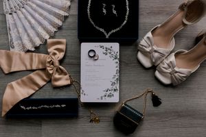 Wedding accessories including fan, jewelry box, shoes, and invitation arranged on rustic wooden surface.