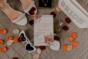 Wedding details flatlay with invitation card, sandals, rose petals, heart sunglasses and bride text on quilted background.