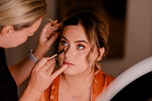 A makeup artist applies cosmetics to a model wearing an orange robe during a professional beauty session.