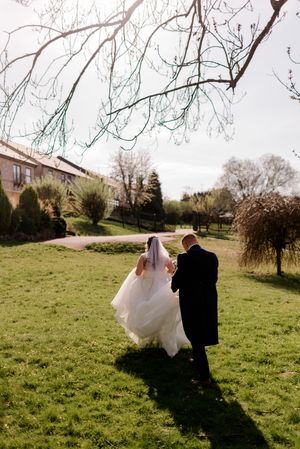 Couple walks across sunlit green lawn in wedding attire as bride's white dress flows behind them on a spring day.