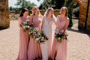Four people in pink and white formal dresses share a candid laughing moment while holding floral bouquets at an outdoor venue.