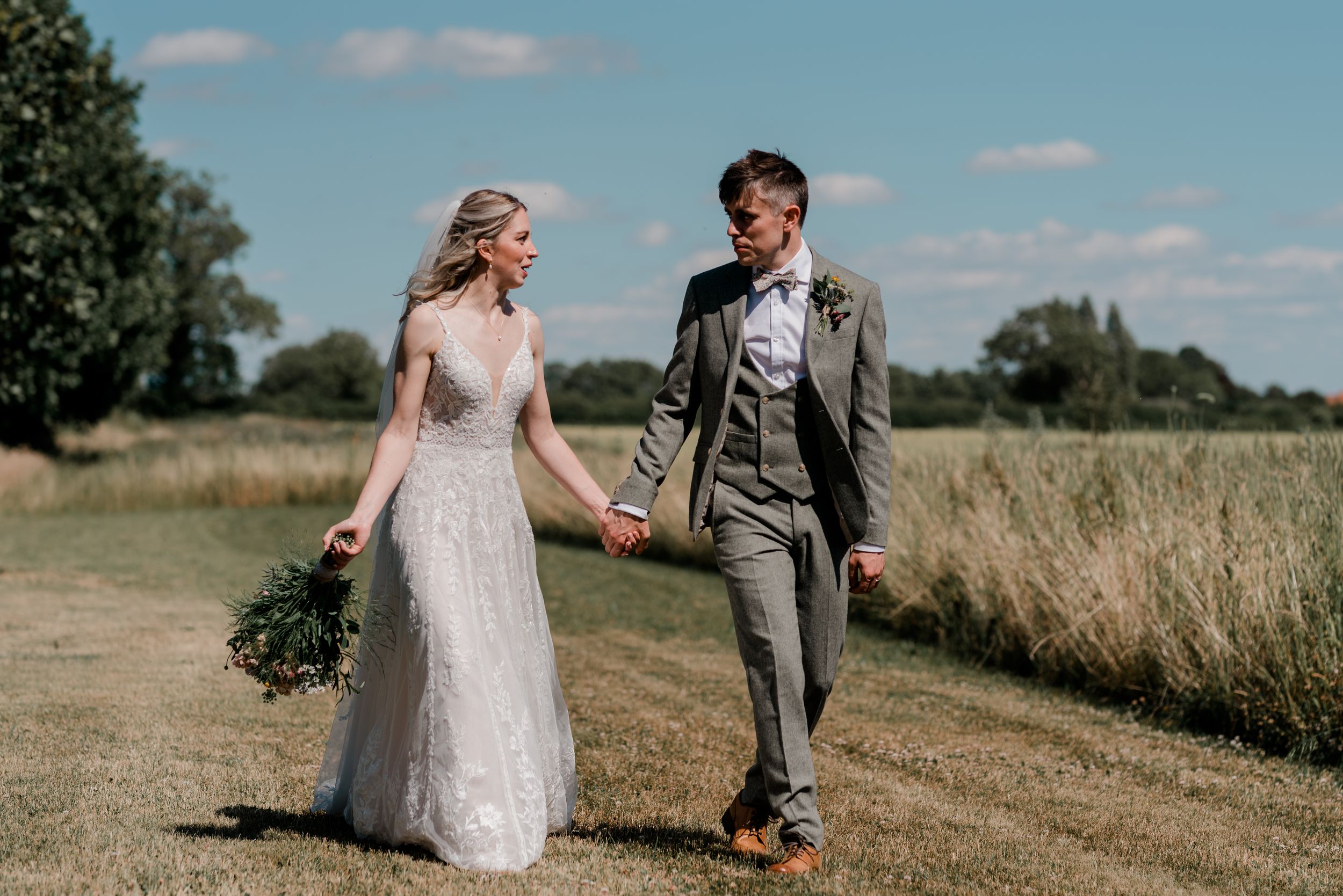 A couple holds hands while walking along a countryside path on their wedding day in summer.