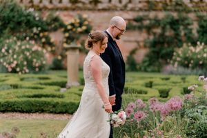 Wedding couple walks through a beautiful garden with pink flowers and manicured hedges beside a historic castle wall.