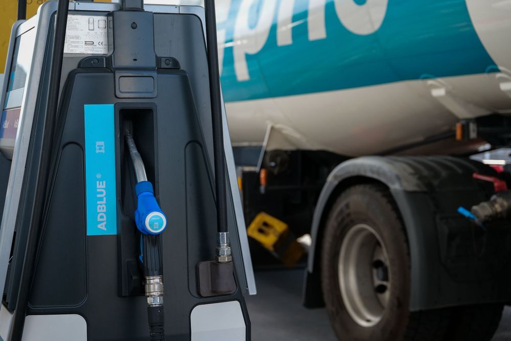 Close-up of AdBlue diesel exhaust fluid dispenser nozzle at a Prio fuel station with tanker in background.