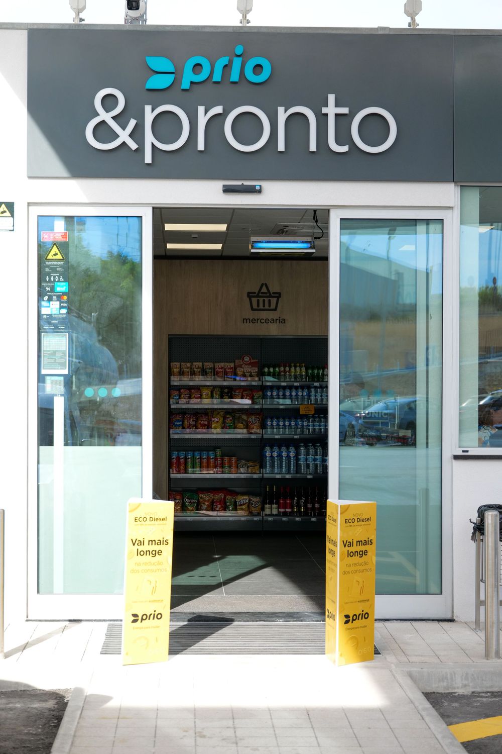 Prio and Pronto convenience store entrance with yellow promotional bollards and shelved products visible inside.