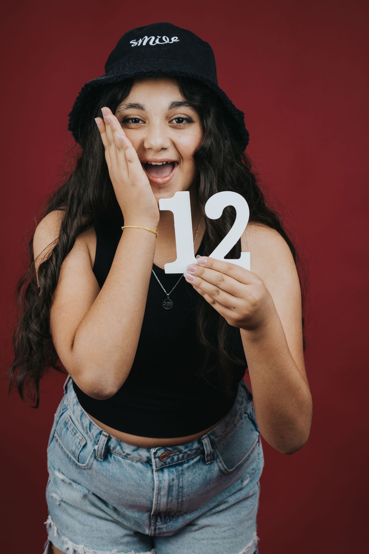 Excited person in black bucket hat and crop top holds up number 12 against red background.