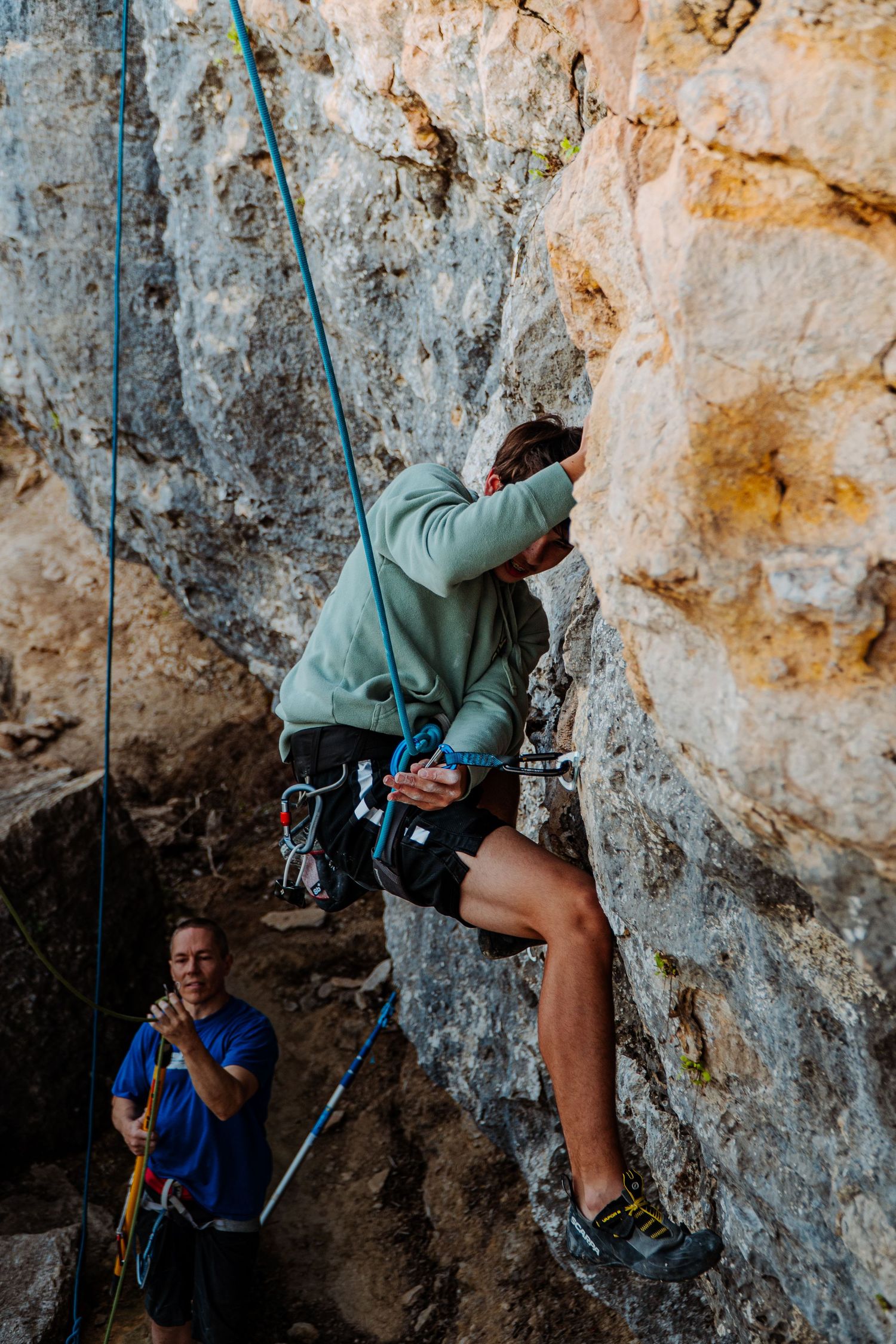 A rock climber ascends a limestone cliff while being spotted by a belayer below.
